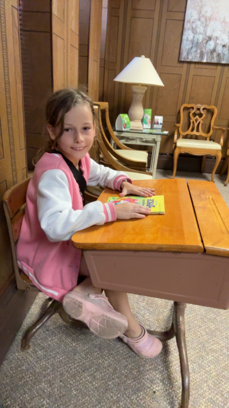 Girl seated at a vintage school desk, wearing a pink jacket. She smiles, looking at the camera.
