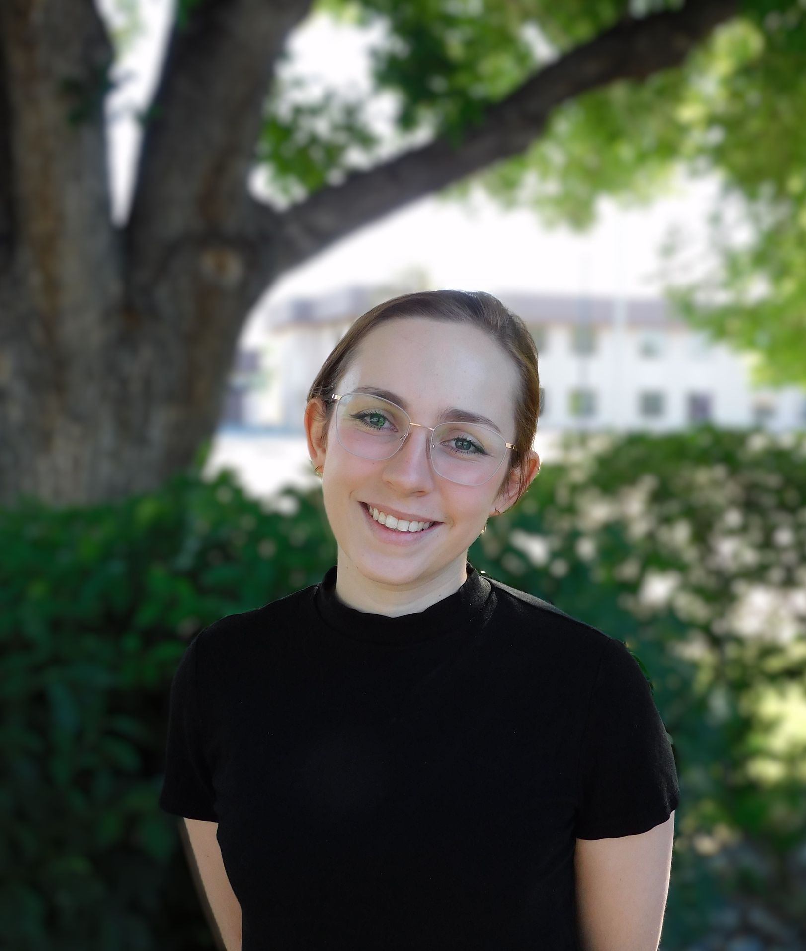 Una mujer joven que lleva gafas y una camisa negra sonríe delante de un árbol.