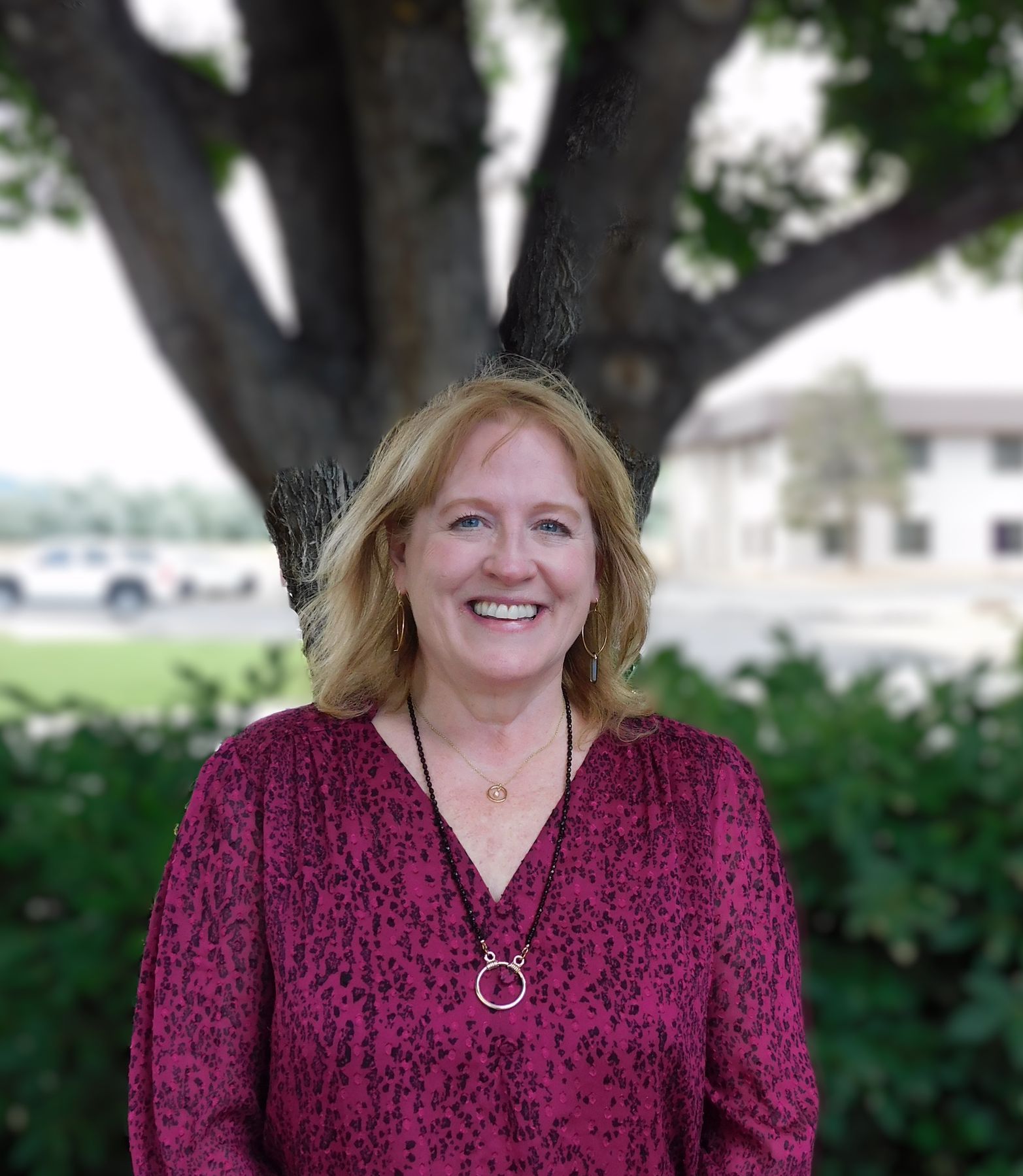 Una mujer con una camisa roja y un collar sonríe frente a un árbol.