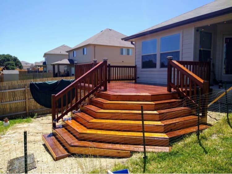 Wooden deck with several steps, stained in a reddish-brown color, next to a house, on a sunny day.