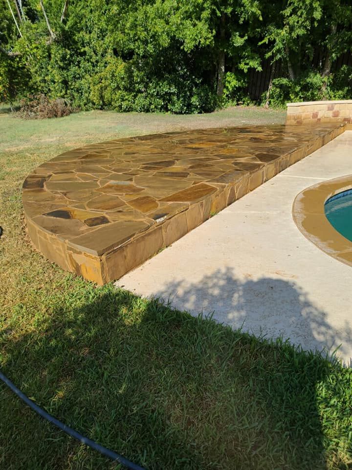 Stone patio with curved edge next to a pool. Brown stones, concrete, green grass, and trees.