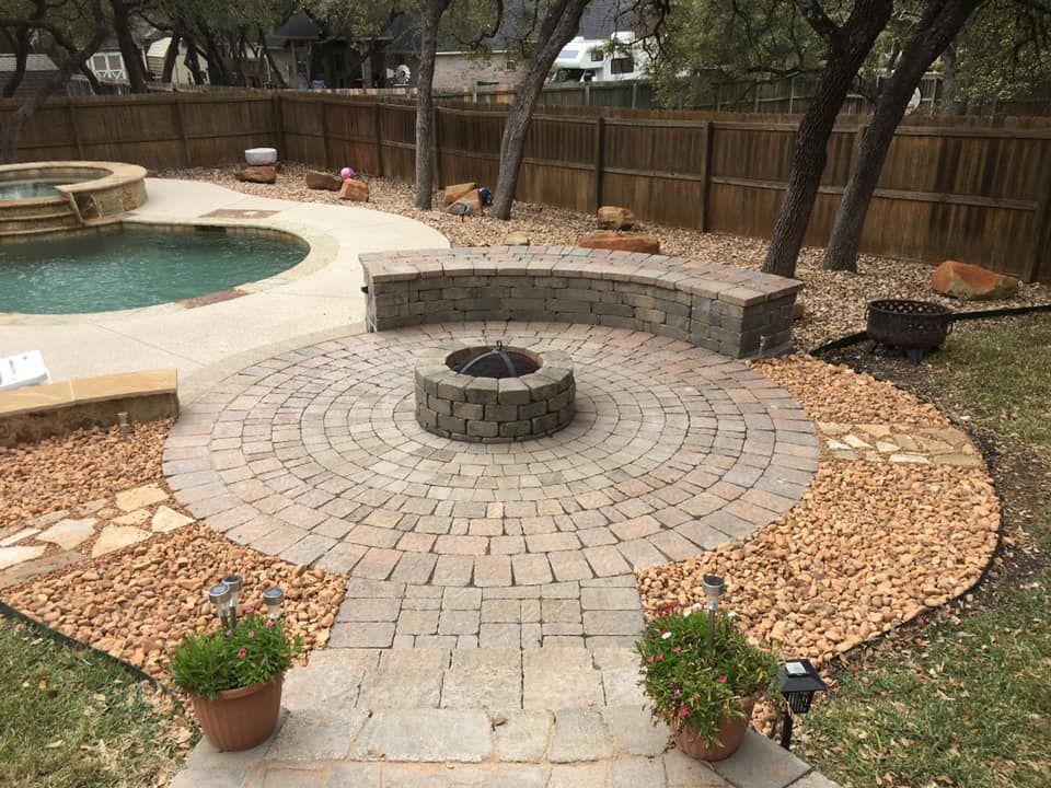 Stone patio with fire pit, seating, and pool in a backyard.