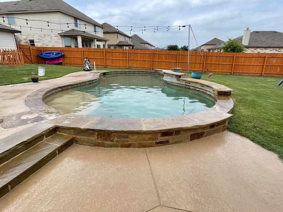 Backyard pool with stone coping and concrete deck, surrounded by a wooden fence and houses.