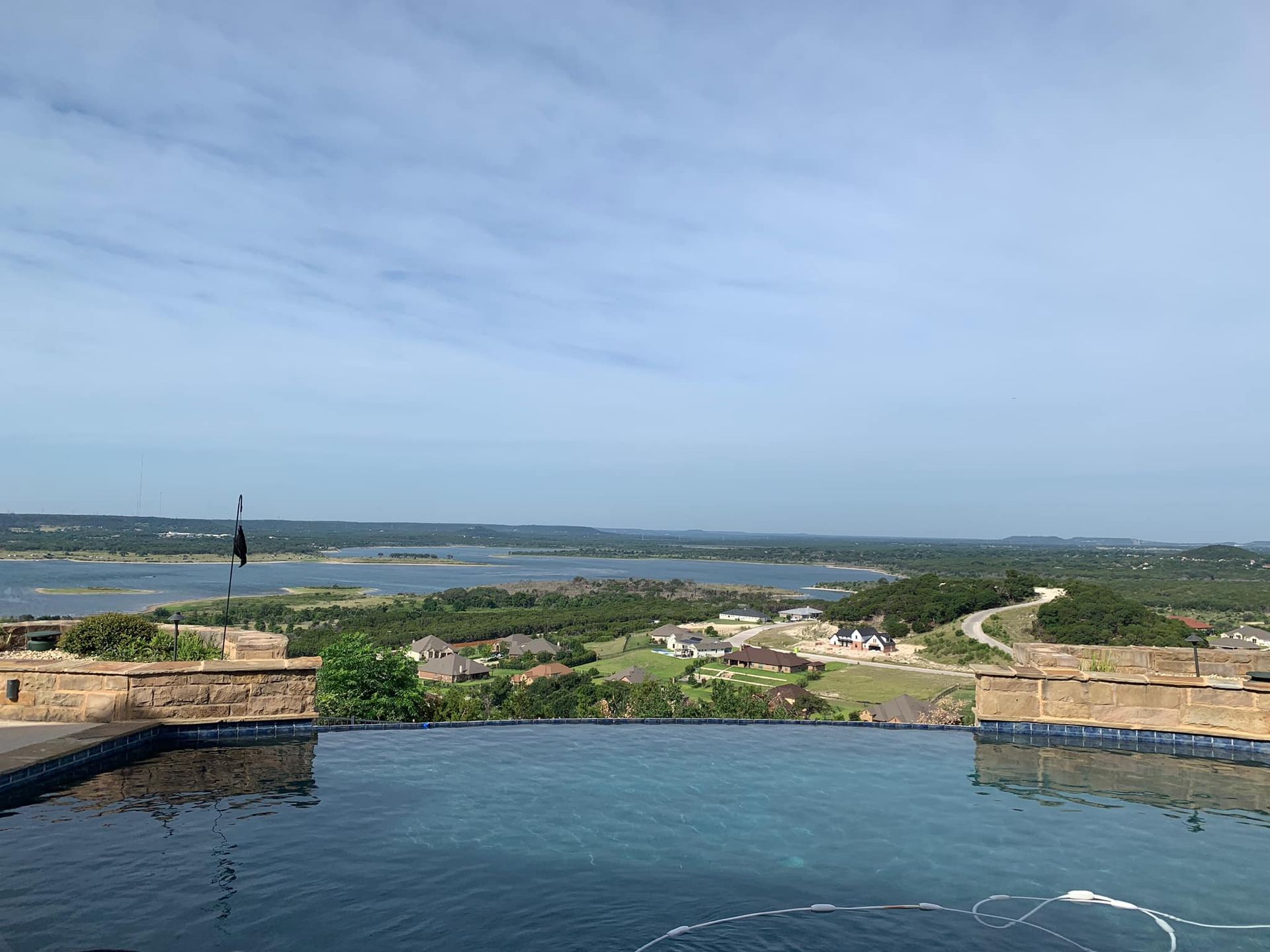 Infinity pool overlooking a lake and green landscape under a partly cloudy sky.