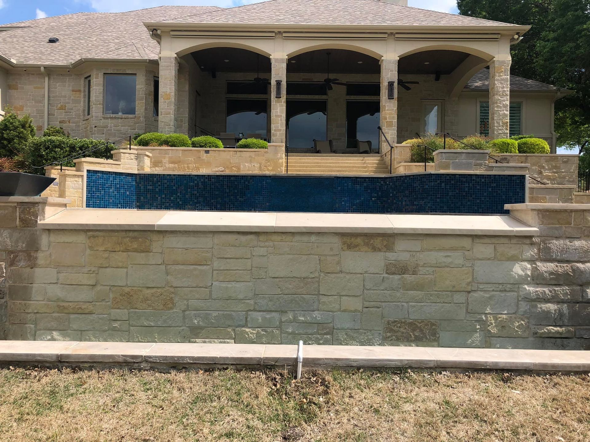 Backyard with a dark blue tile pool overlooking a house with stone walls and a covered patio.