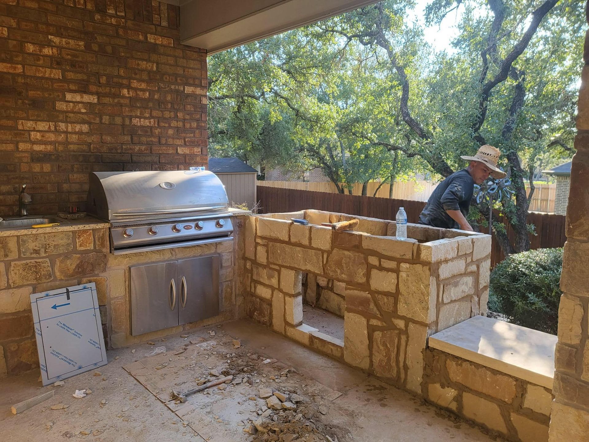 Outdoor kitchen construction. A person works on stone features. A grill and brick wall are visible.