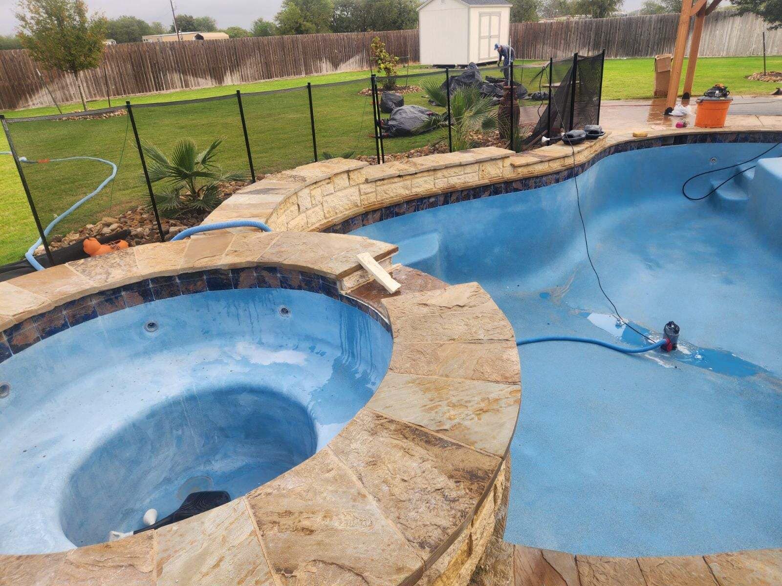 Pool and spa with blue interior, stone coping, black fencing, and a grassy backyard.