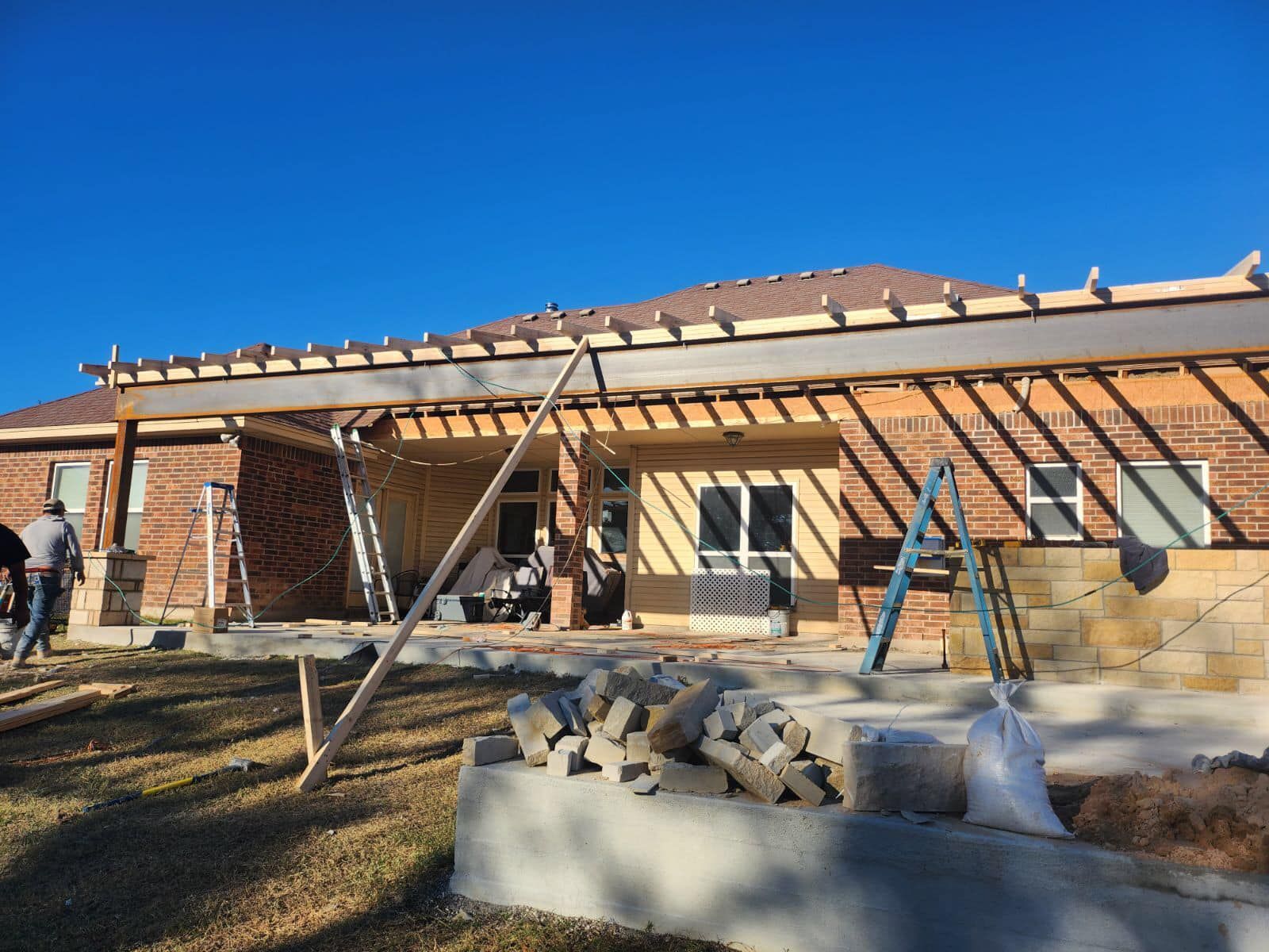 Construction of a patio cover on a brick house. Wood framing is visible; workers are present.