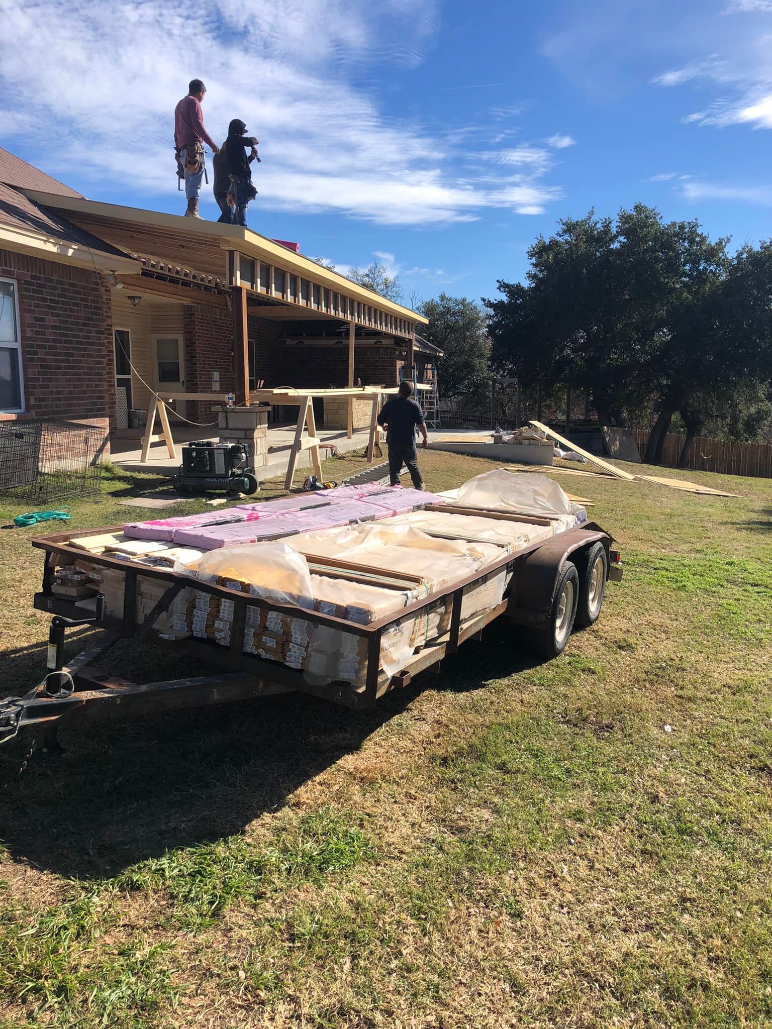 Construction workers on a roof, building a structure. Trailer with insulation on the grass in front of the house.
