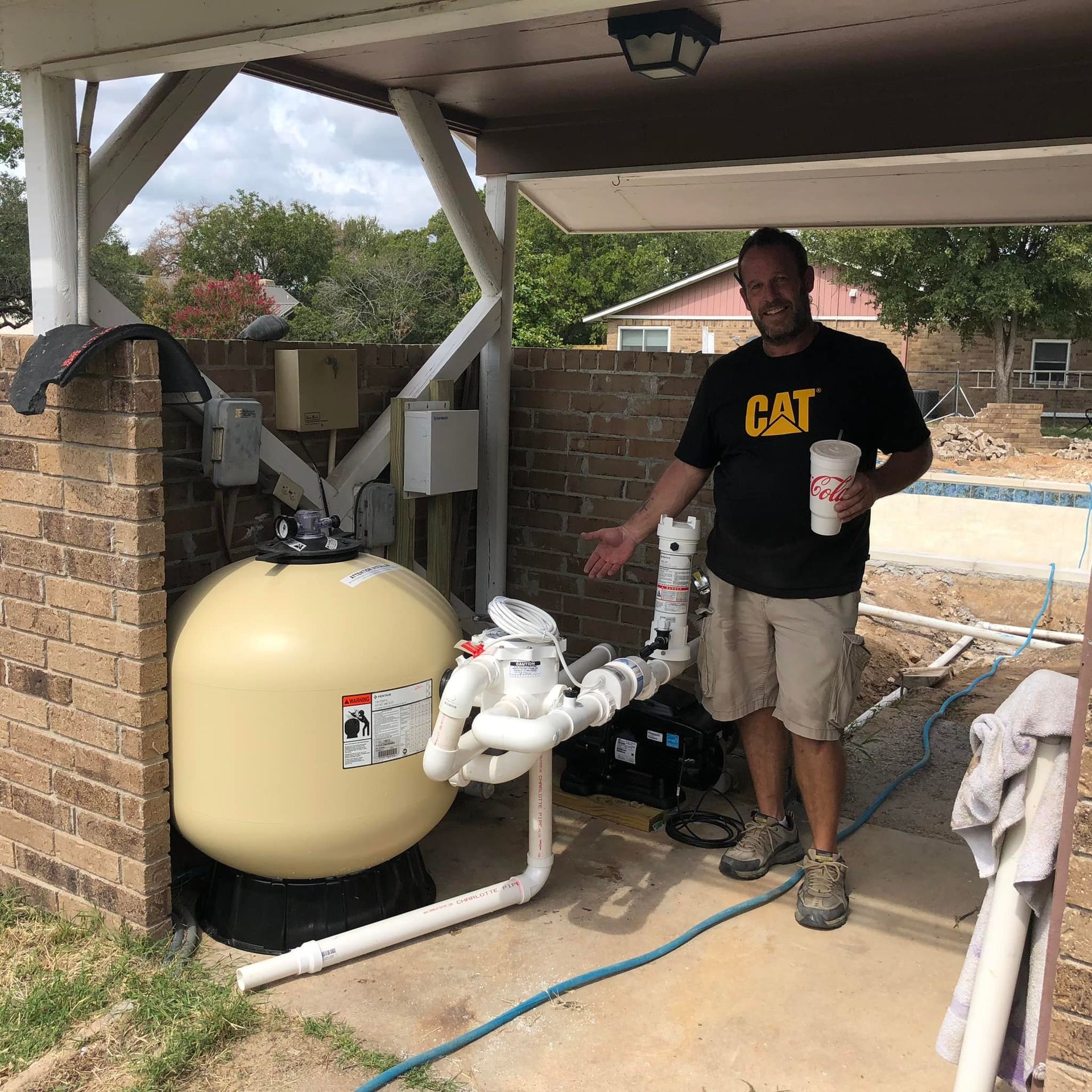 Man pointing at pool filter equipment under a covered area; beige tank, brick wall, and blue hose visible.