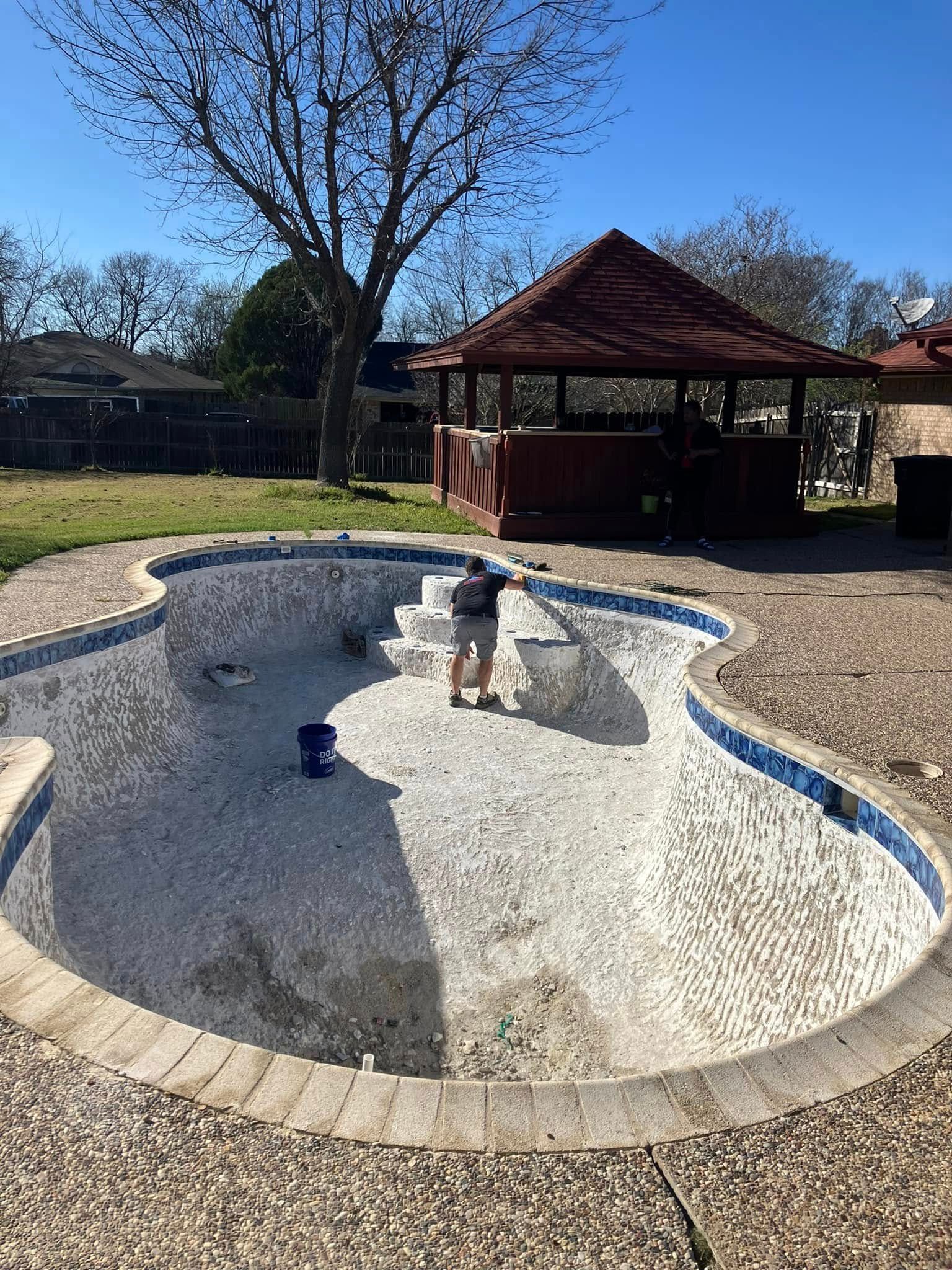 Person cleaning an empty, mosaic-tiled swimming pool on a sunny day with a gazebo in the background.