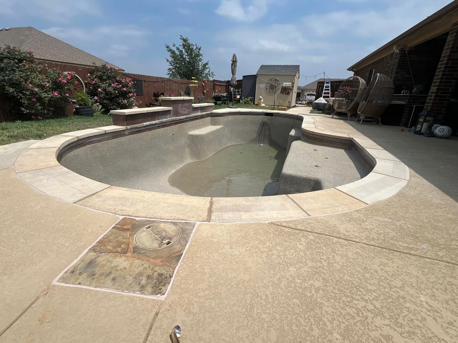 Empty, kidney-shaped swimming pool with stone coping and concrete deck, in a residential backyard on a sunny day.