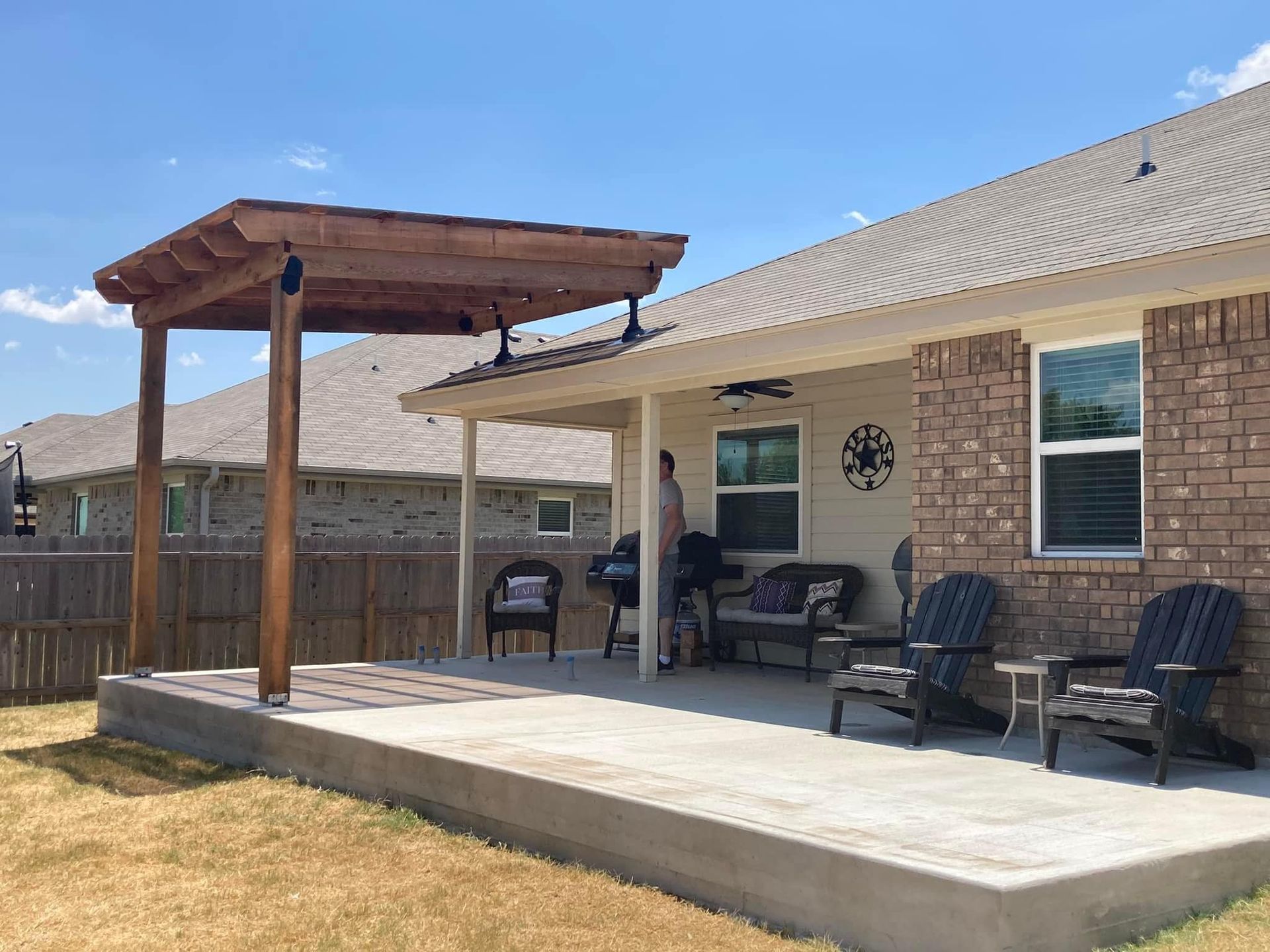 Patio with a pergola, concrete slab, seating, and a person near a grill. Sunny day.