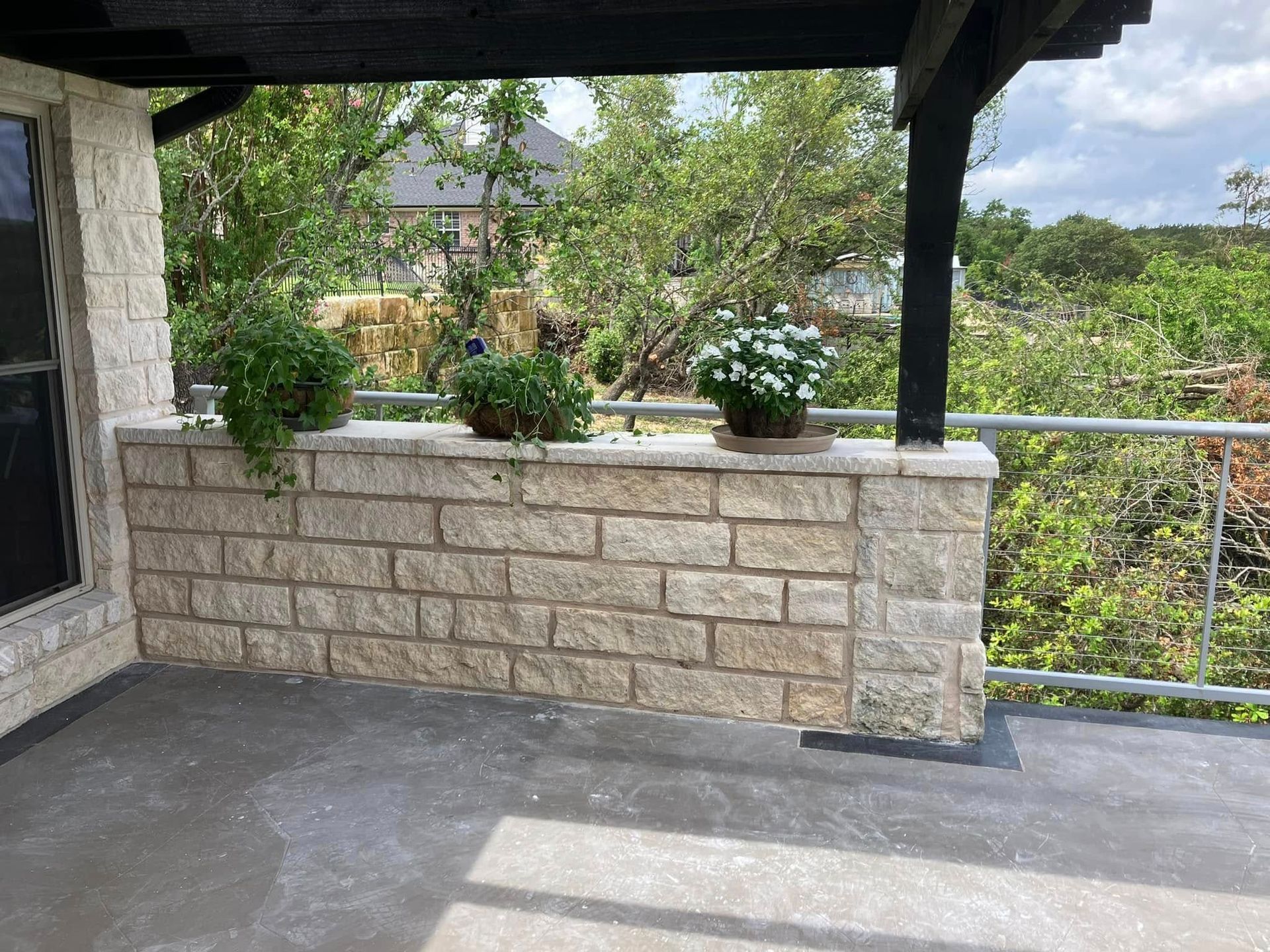 Stone patio with plants on a low wall, under a shaded overhang, with a wooded view.