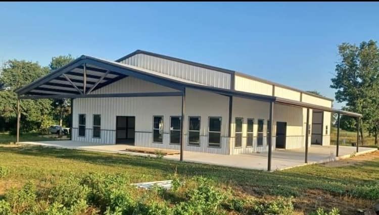 Cream-colored metal building with dark trim and awning, concrete patio, windows, and clear blue sky.