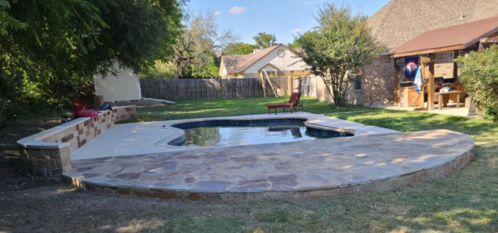 Backyard with a pool surrounded by stone and a covered patio with a flag hanging.