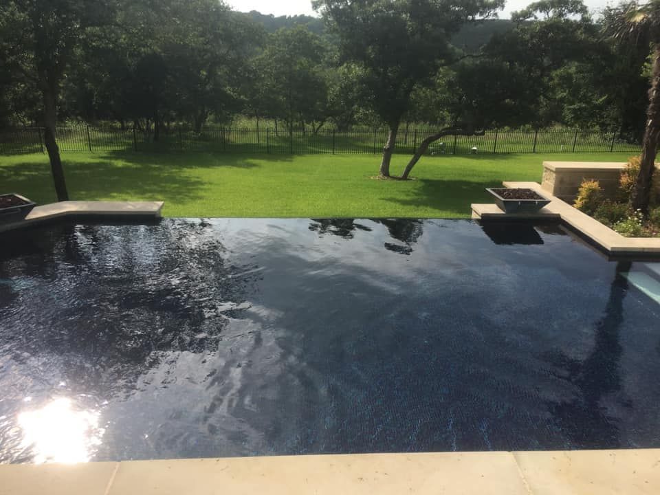 A dark blue tiled infinity pool reflecting the sky, set in a grassy yard with trees in the background.