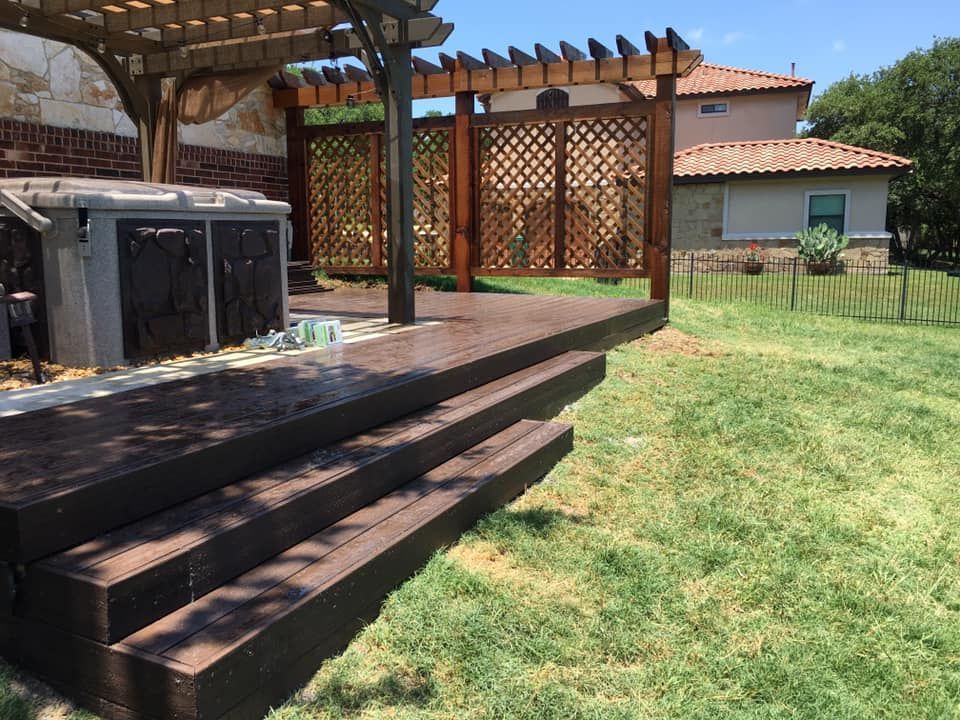 Brown wooden deck and steps next to a hot tub under a pergola in a backyard.