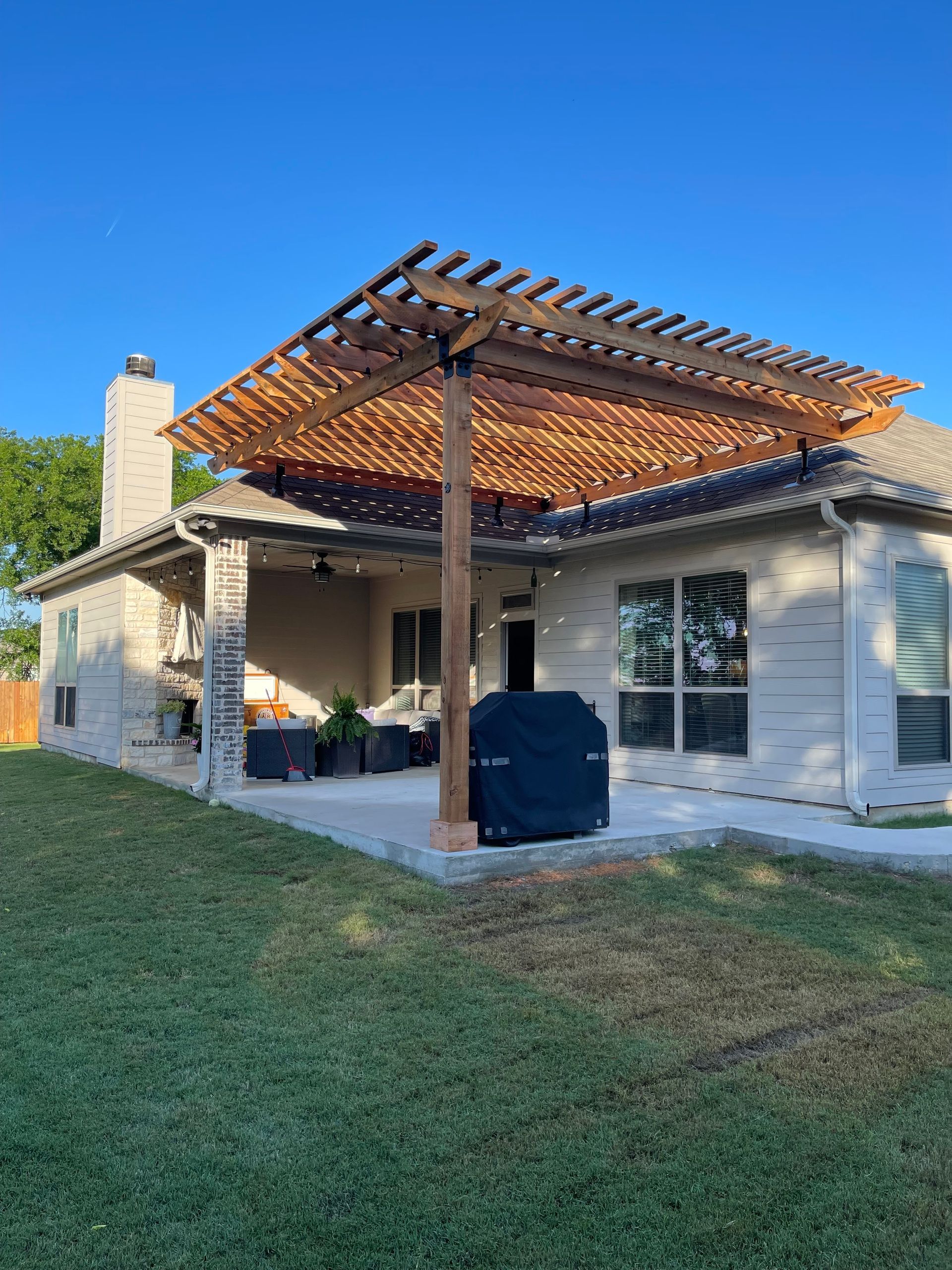 Wooden pergola over a patio, attached to a brick house. Includes a grill and a grassy yard.