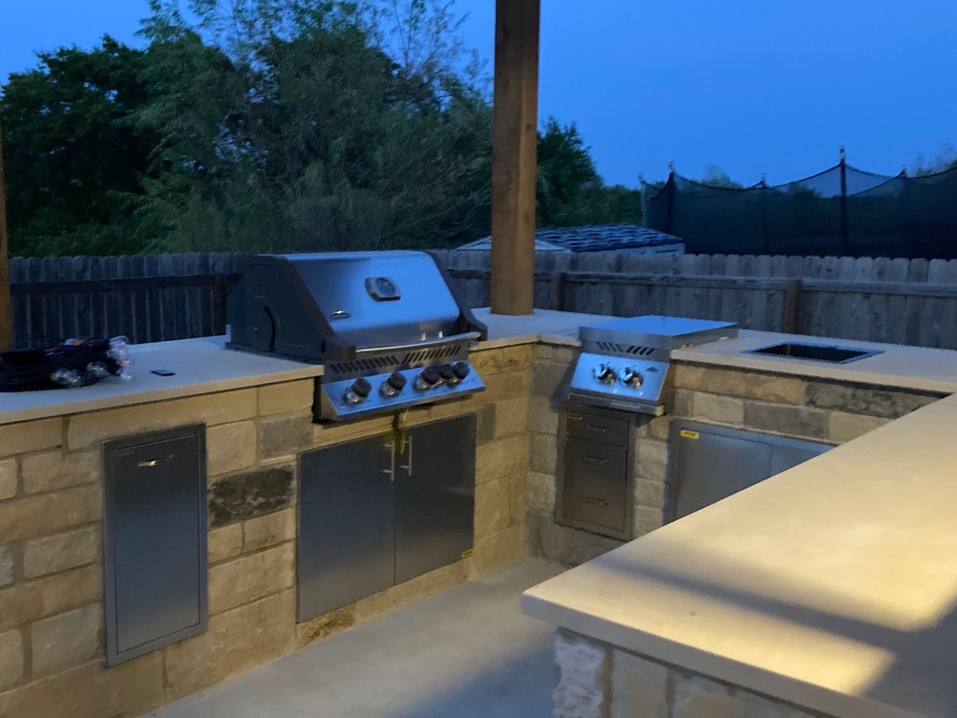 Outdoor kitchen with built-in grills, cabinets, and counter space under a covered patio. Dusk lighting.