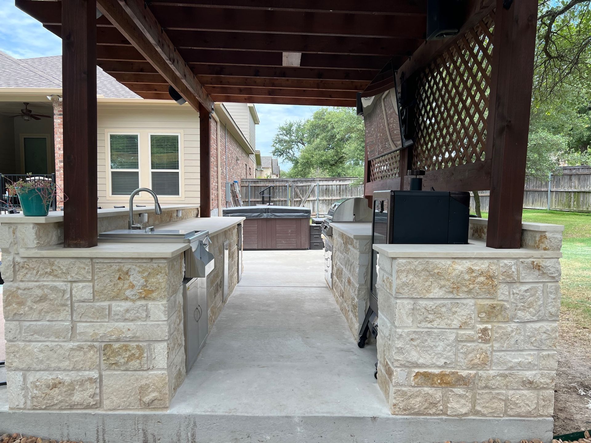 Outdoor kitchen with stone counters, brown pergola, grill, sink, and spa in the background.
