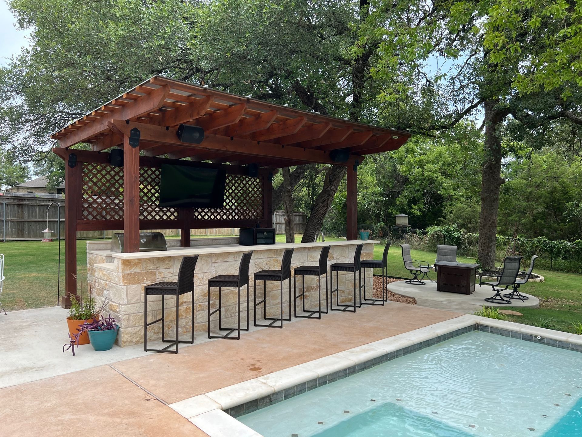 Outdoor bar with a TV, stools, and pergola near a pool.