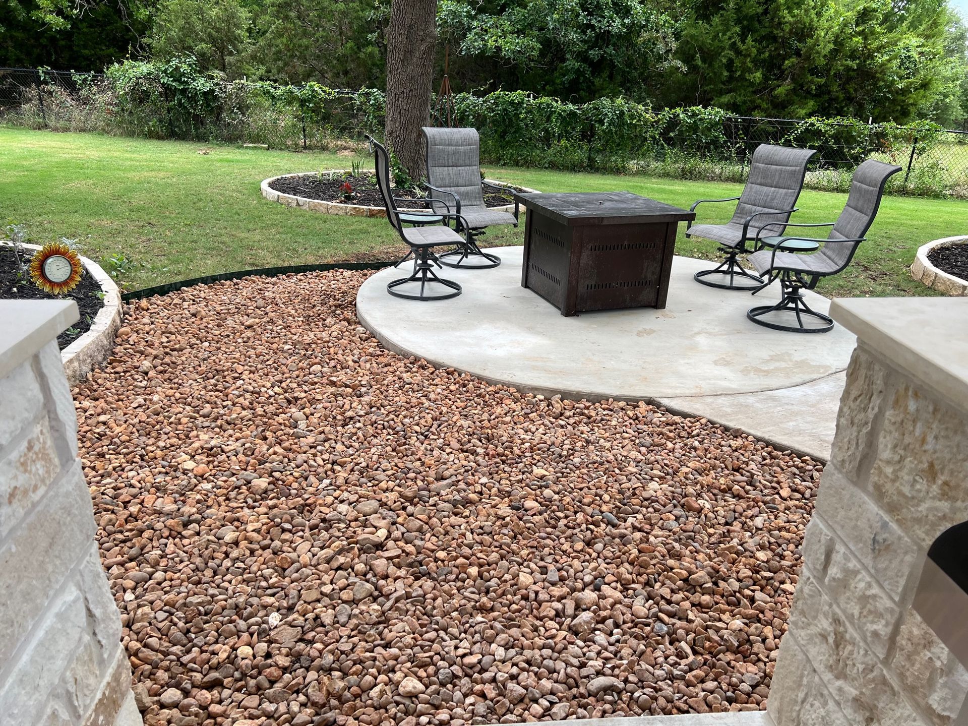 A backyard patio with a fire pit, seating, and a gravel pathway. Green grass and trees are in the background.