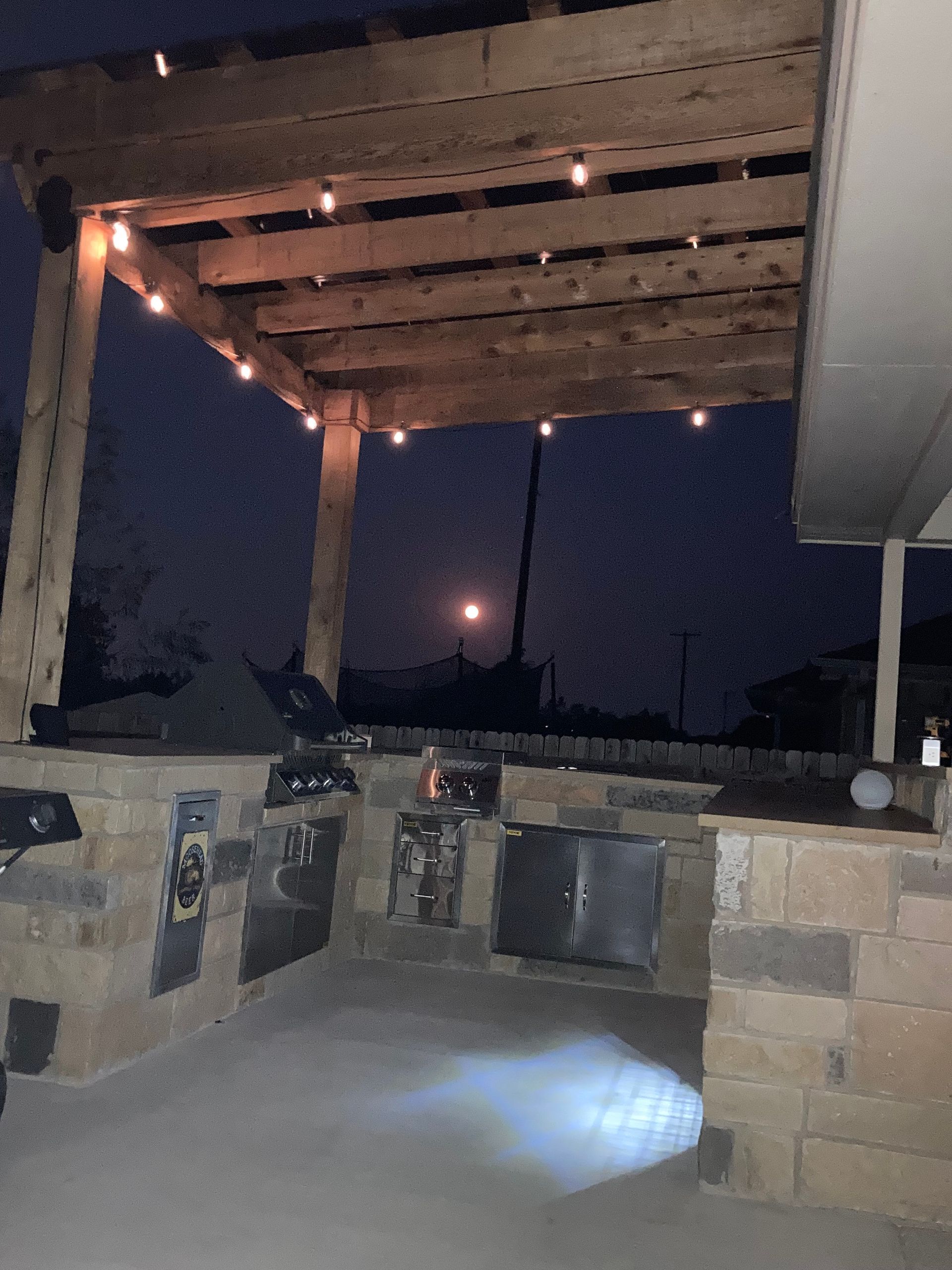Outdoor kitchen area with wooden pergola, string lights, stone walls, and grills at night.