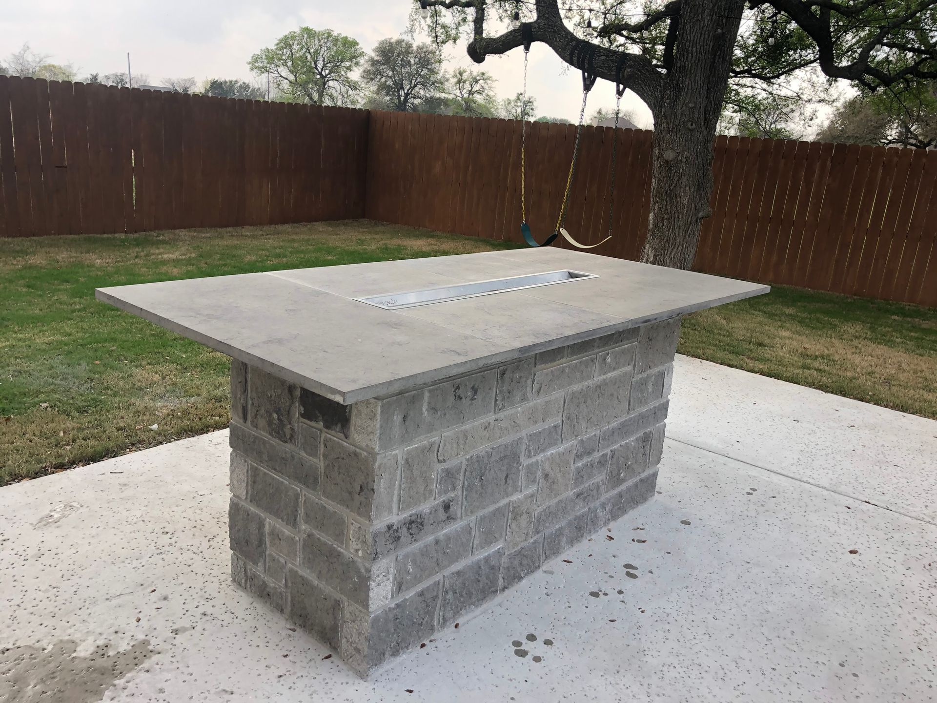 Stone fire pit on a concrete patio in a backyard with a wooden fence.