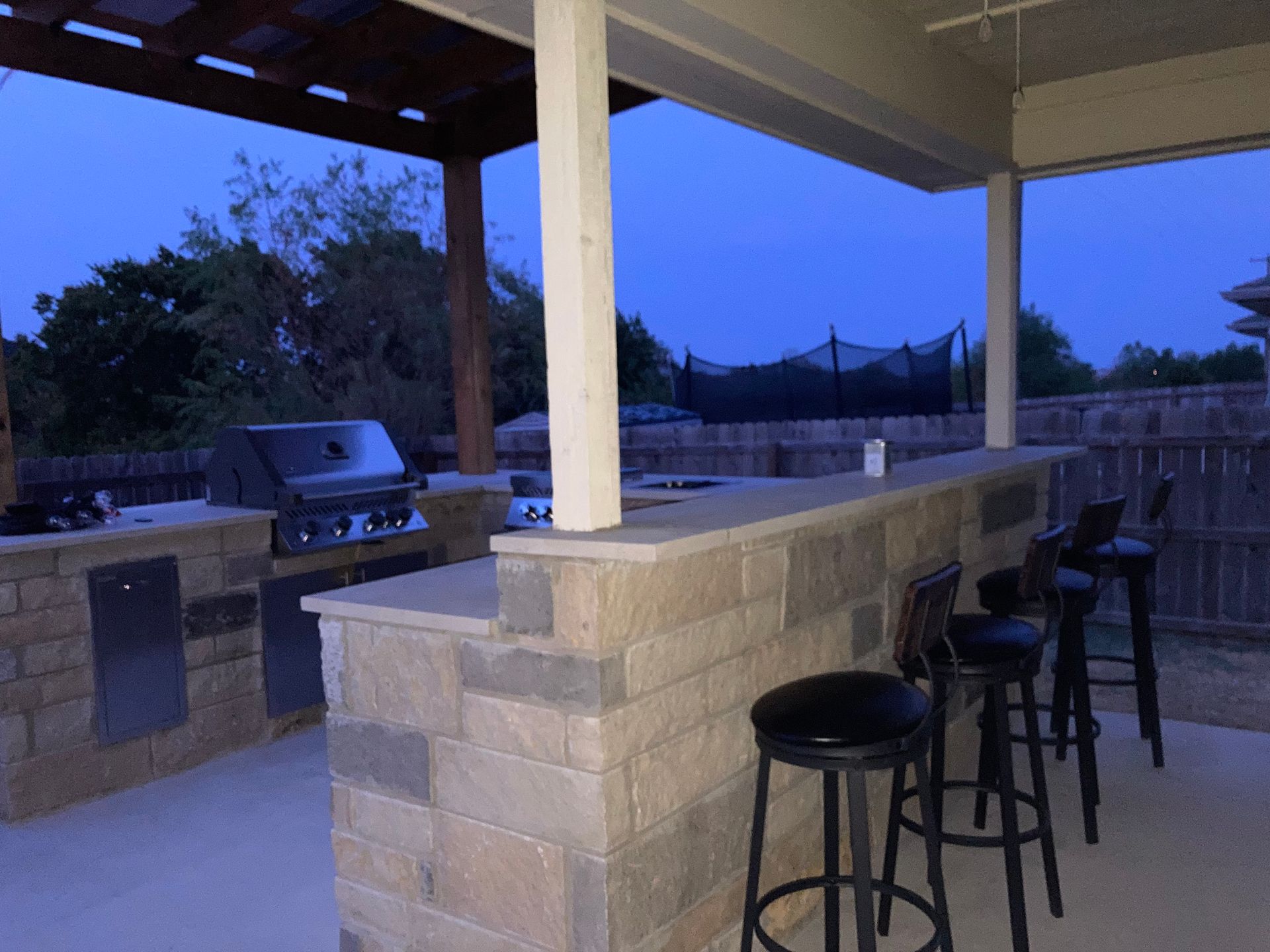 Outdoor kitchen with grill, bar, and seating, under a covered patio at dusk.