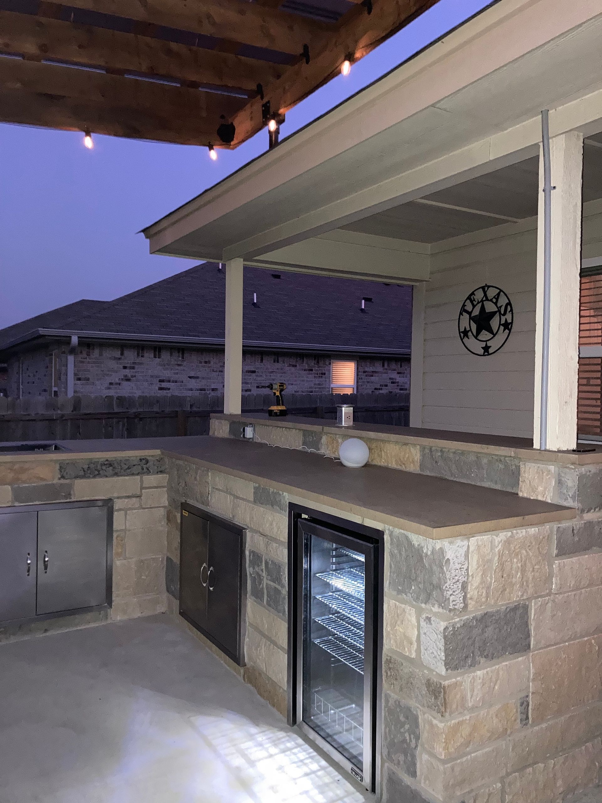 Outdoor kitchen with stone counters, a grill, and a beverage cooler, under a covered patio.