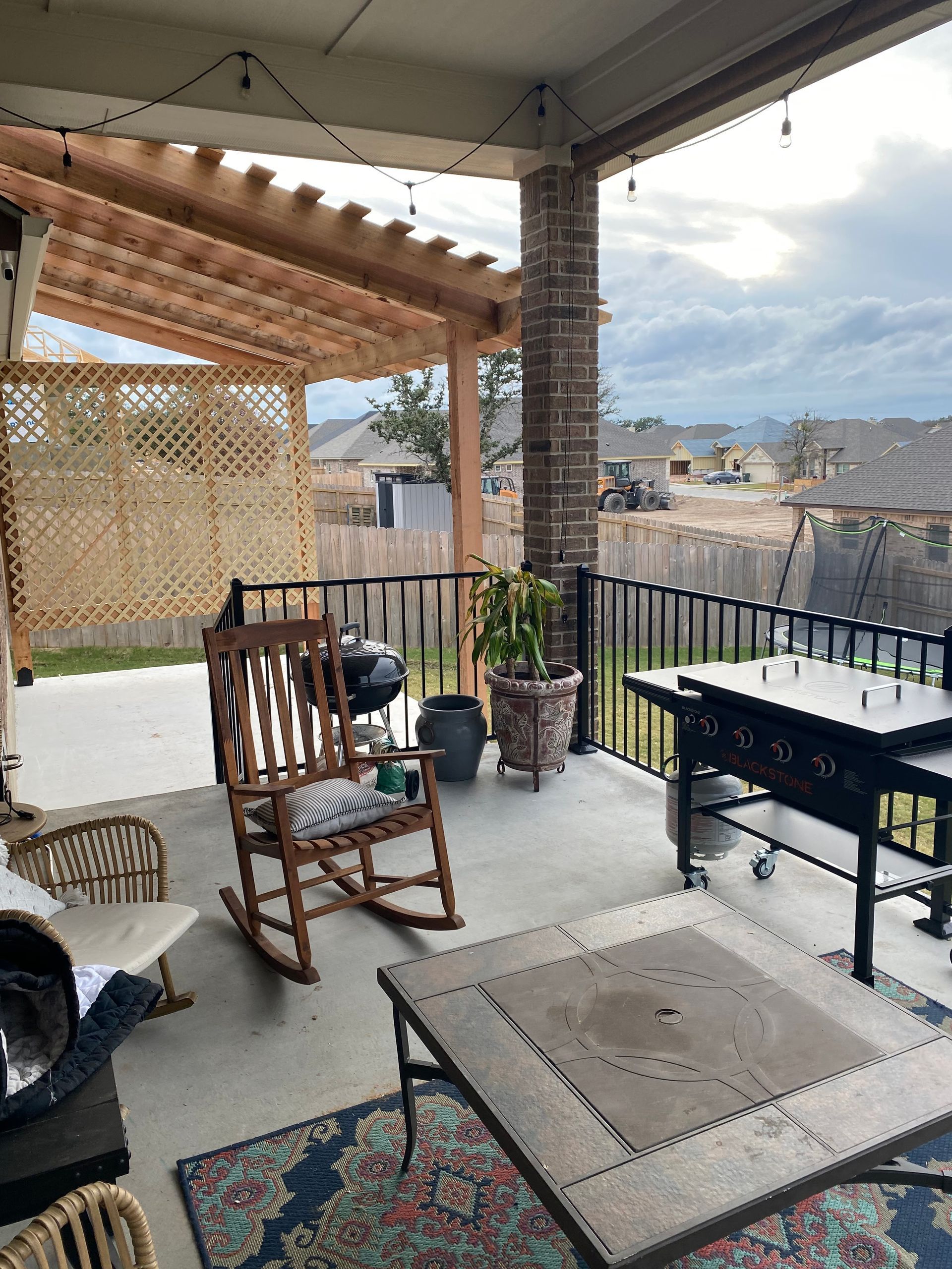 Covered patio with rocking chair, grill, and outdoor furniture. Backyard view with cloudy sky.