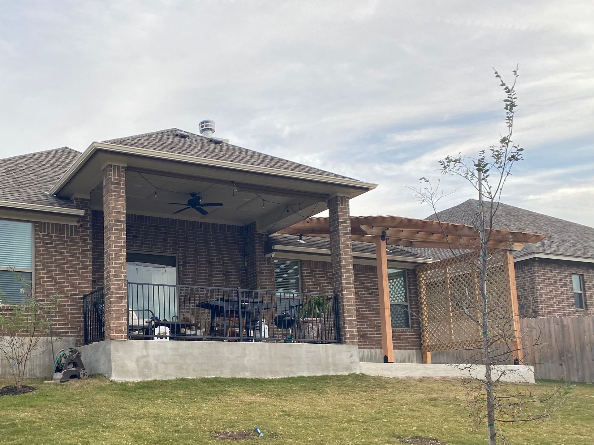 Brick house with a covered patio and pergola in a grassy yard under a cloudy sky.