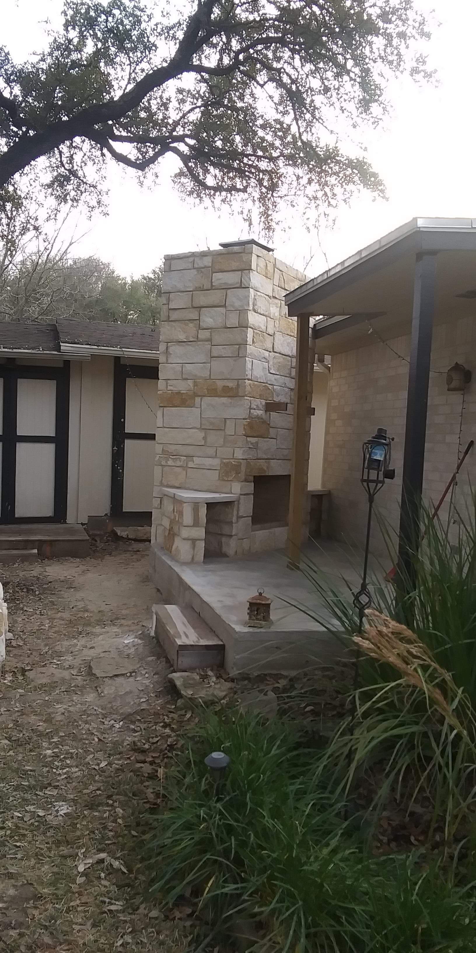 Stone chimney on a patio next to a house with an overgrown yard.