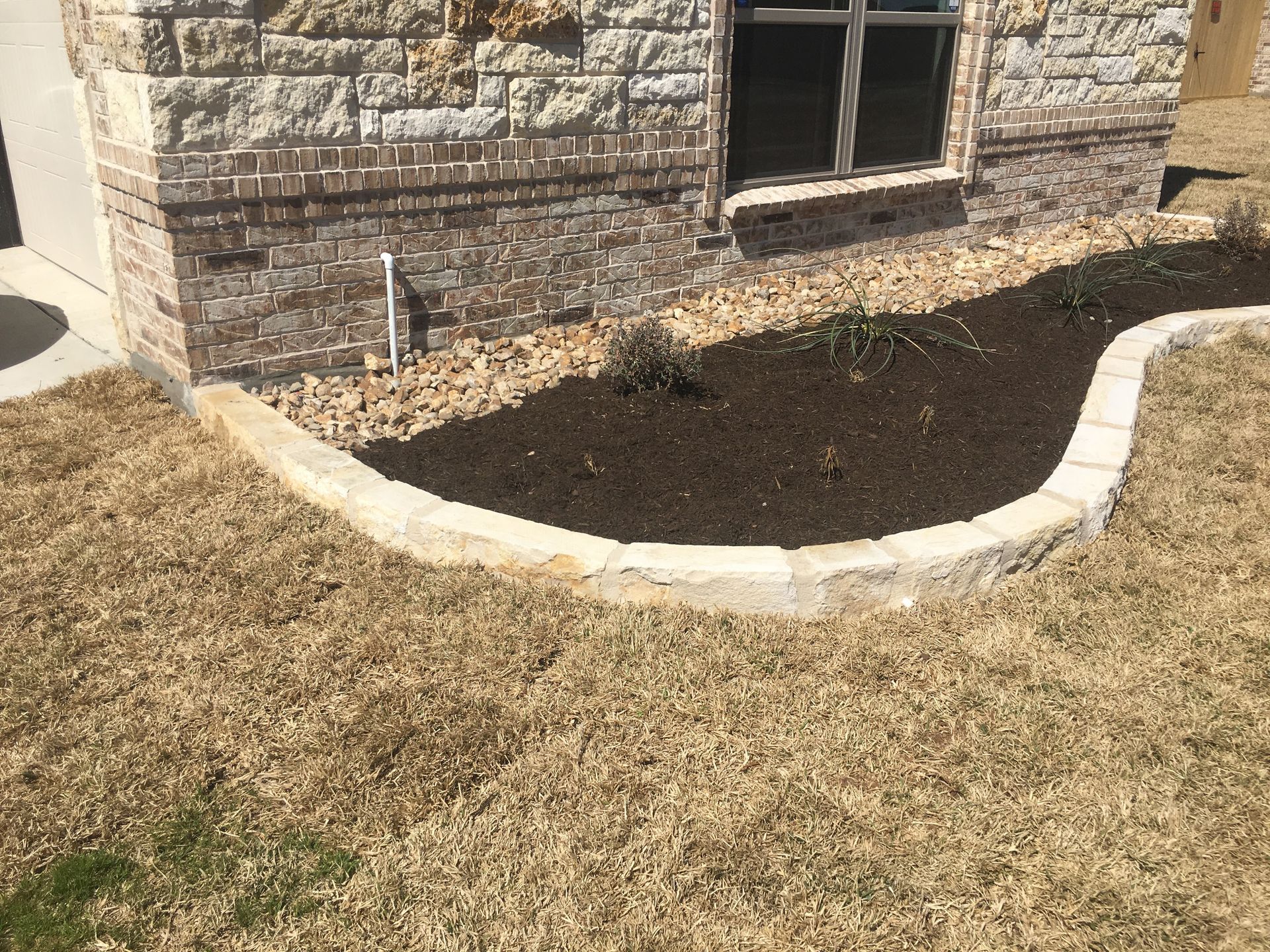 Flower bed with stone border, mulch, and small plants against a brick and stone house. Dry grass in foreground.