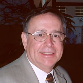 Man with glasses, smiling, in a suit and tie, outdoors with building in background.