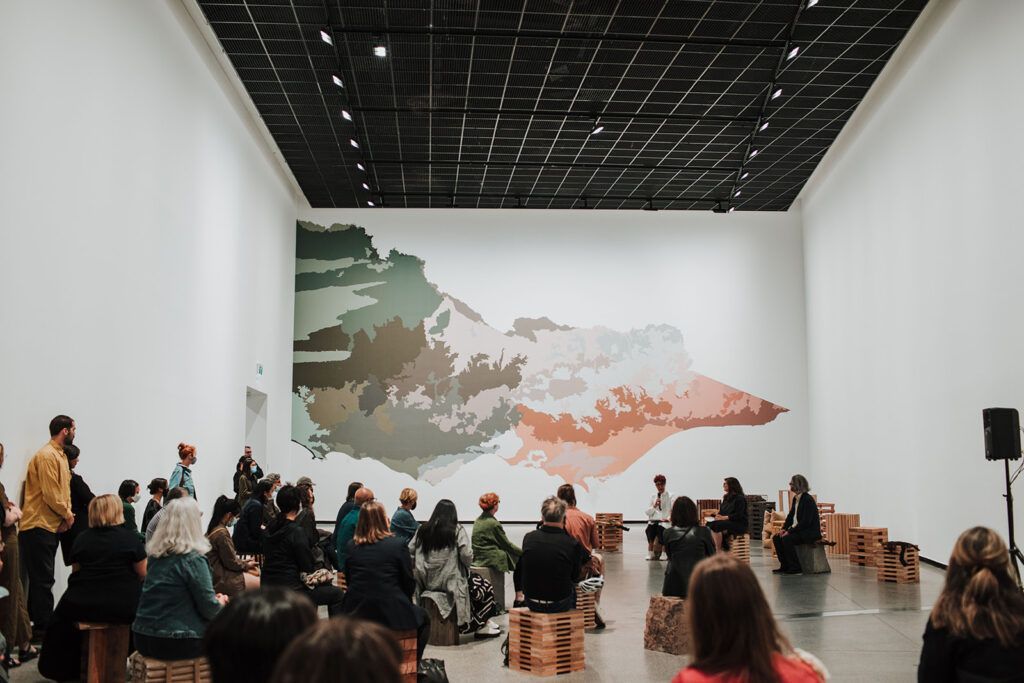 People seated, watching a performance in a gallery; large abstract landscape painting on the wall.