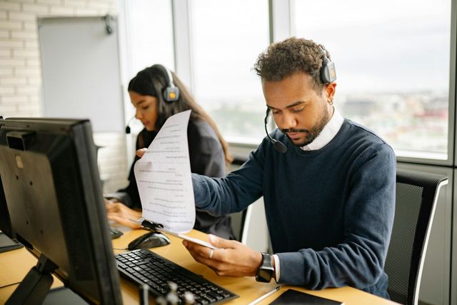 Two people in an office wearing headsets, looking at documents and computer screens.