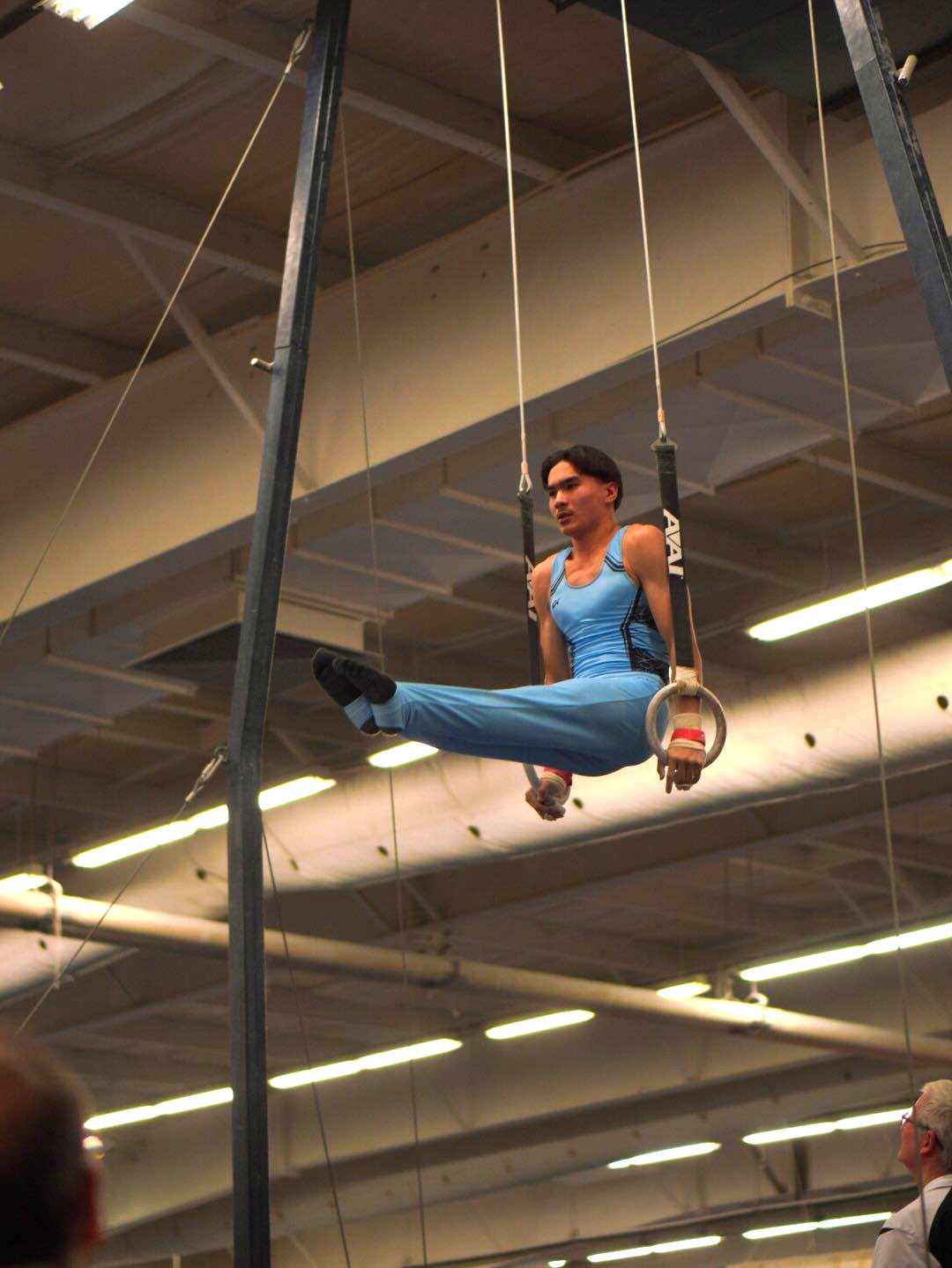 Gymnast performing a straddle planche on rings, wearing a blue leotard in an indoor setting.