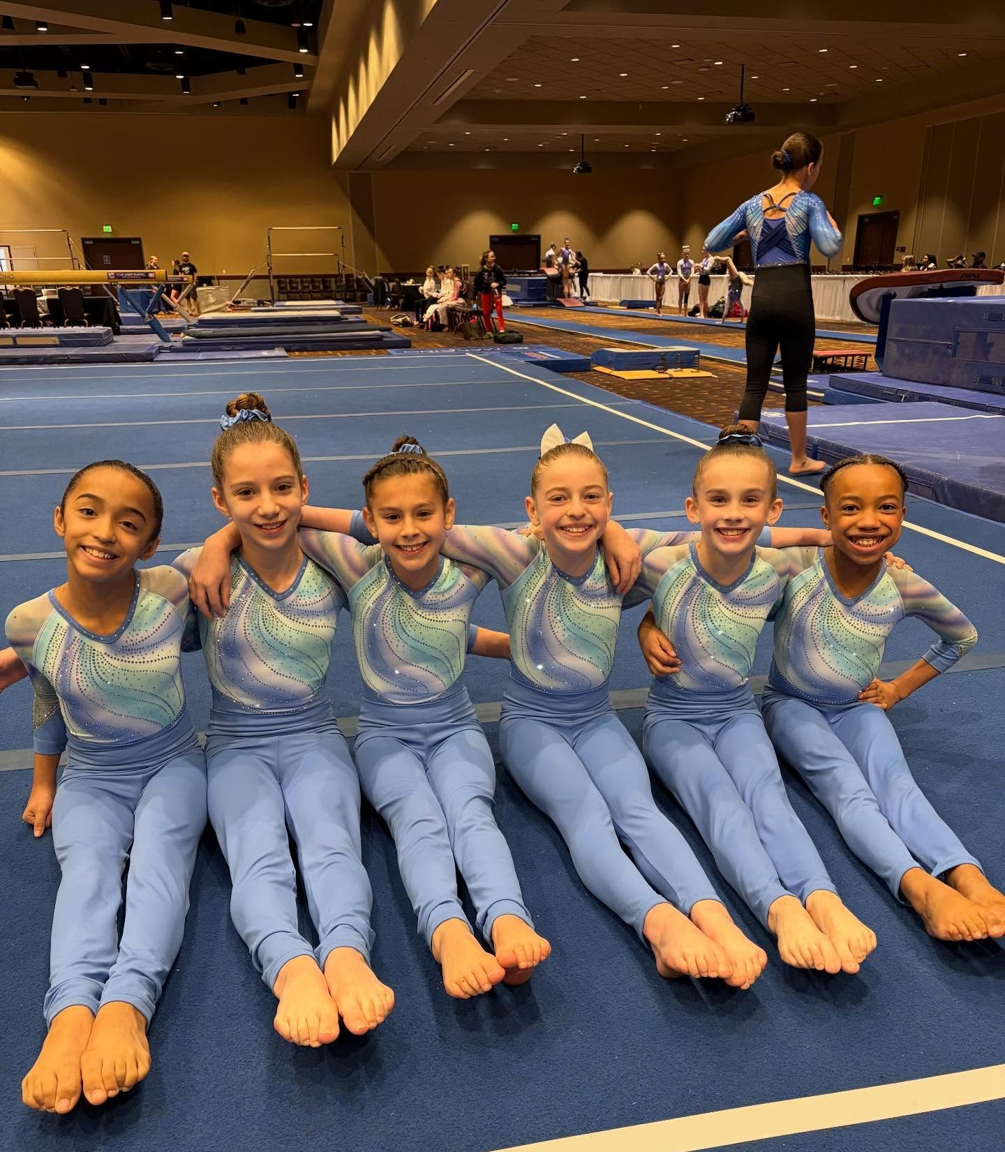 Six Parkettesin blue outfits pose on a blue mat. They are smiling and have their arms around each other.