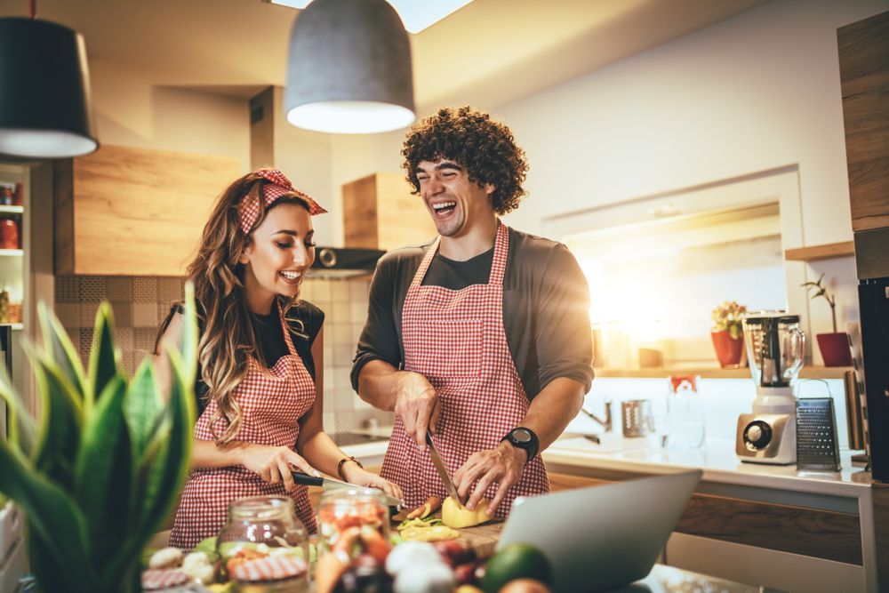 A man and a woman are cooking in a kitchen while looking at a laptop.