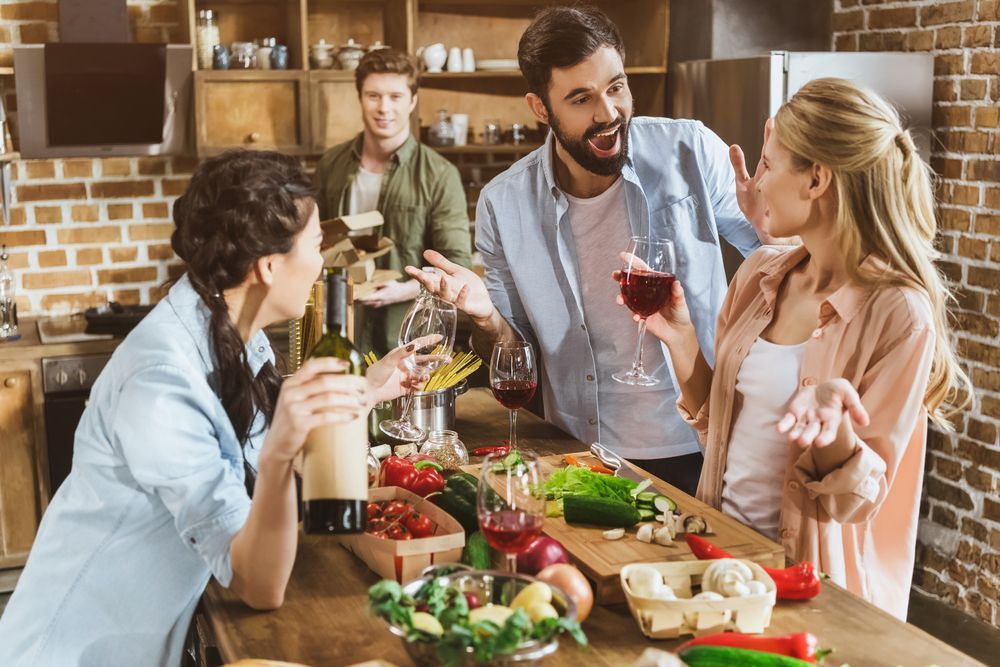 A group of people are standing around a table in a kitchen drinking wine.