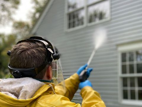 A man in a yellow jacket is cleaning a house with a high pressure washer.