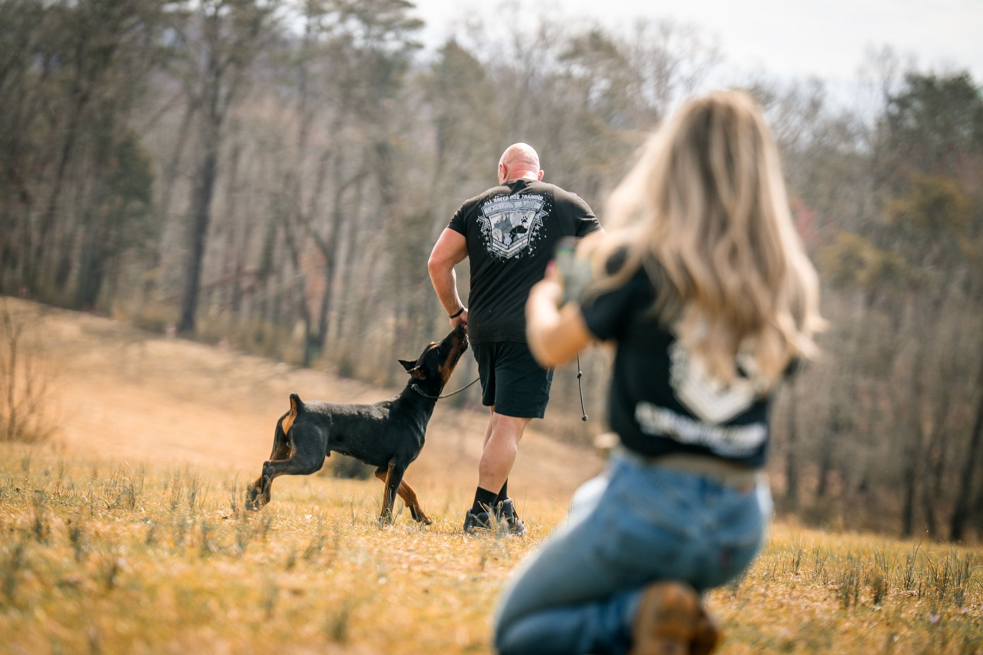A person kneels in a field while photographing a dog jumping toward another person walking in the distance.