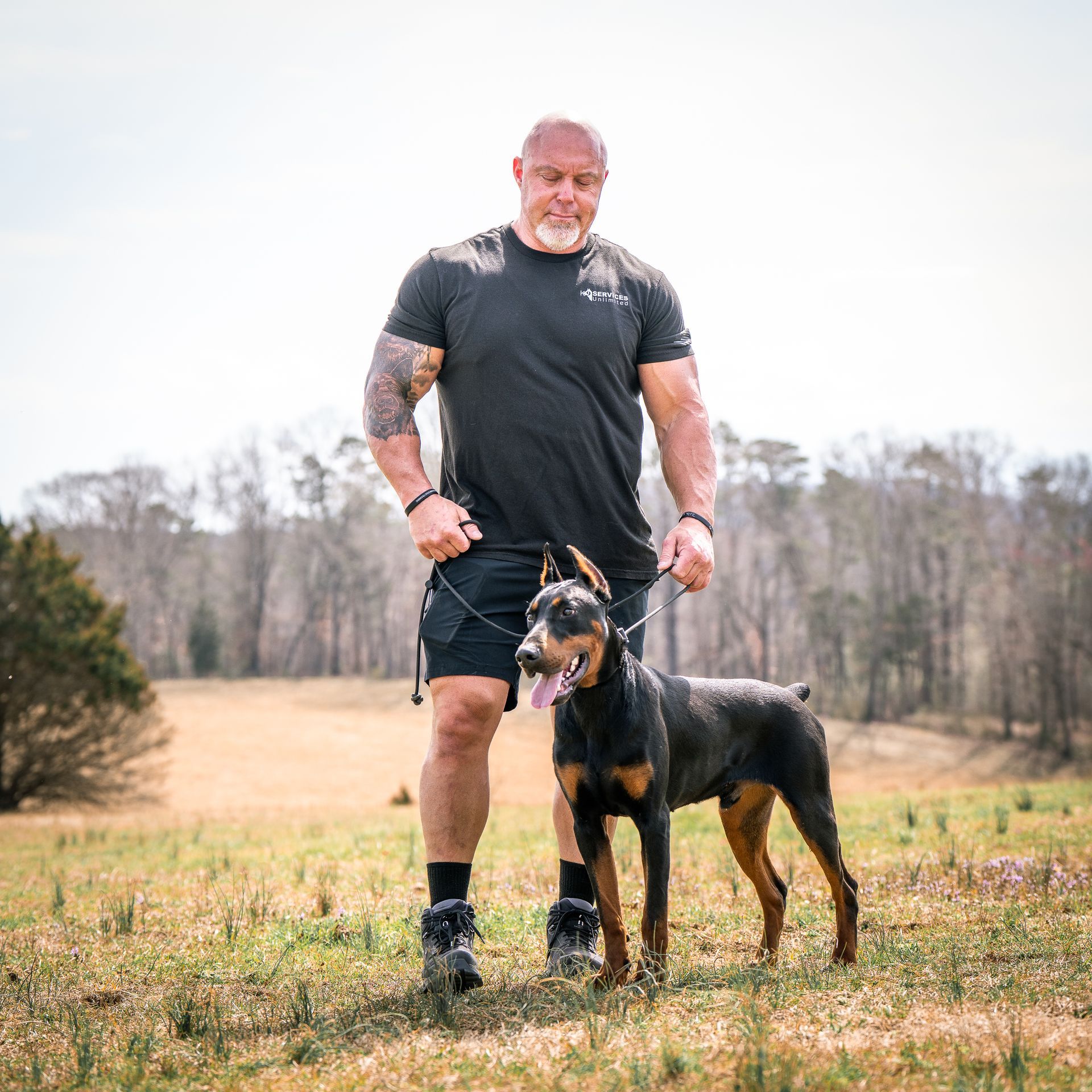 A muscular person in a dark t-shirt stands in a field holding the leash of a black and tan Doberman with its tongue out.