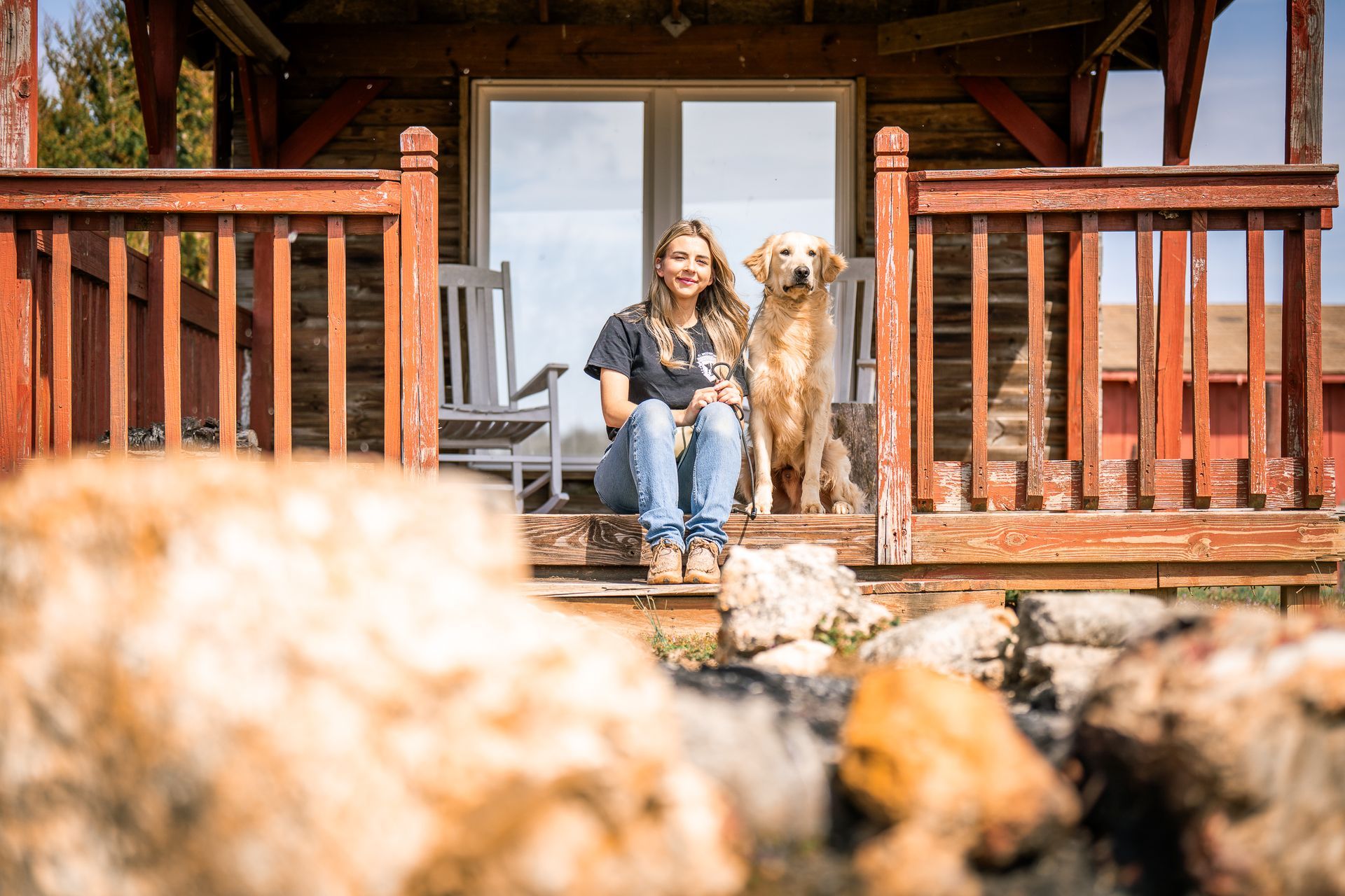 A woman sits on a wooden cabin porch next to a golden dog, with out-of-focus rocks in the foreground.