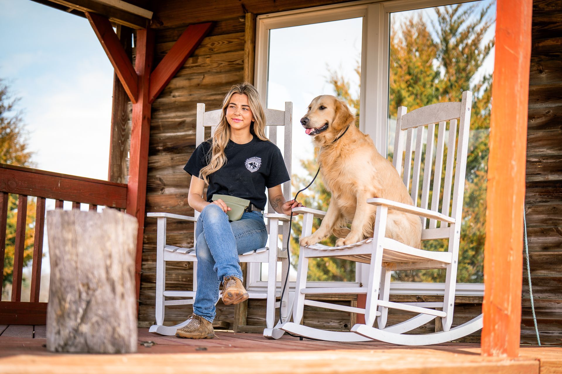 A person in a black t-shirt and jeans sits on a white rocking chair on a wooden porch with a golden retriever beside them.