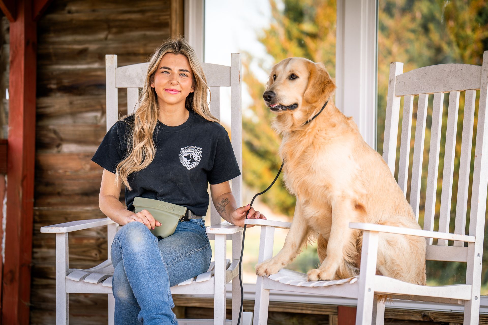 A person with long blonde hair sits in a white wooden rocking chair with a golden retriever, holding its leash.