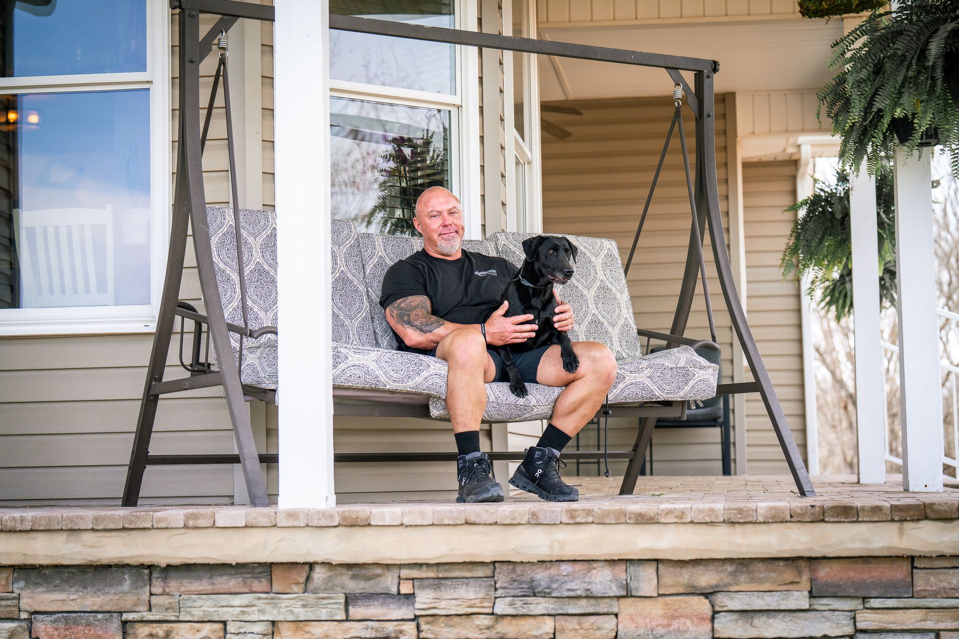 A person sits on a porch swing with a black dog on their lap, smiling in front of a house.