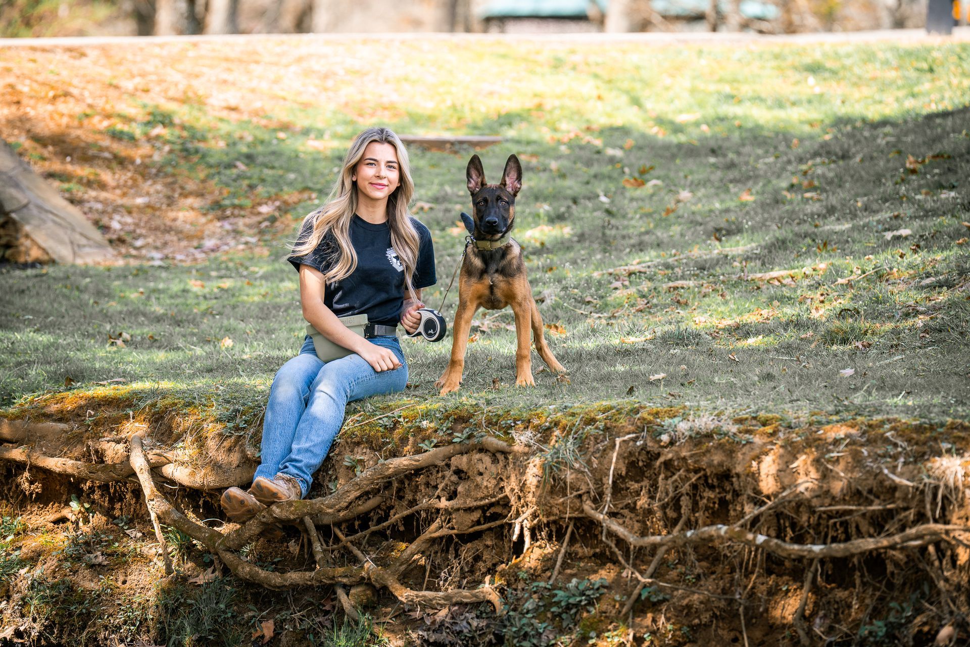 A person and a German Shepherd dog sit together on a grassy, earth-banked edge in an outdoor, wooded setting.